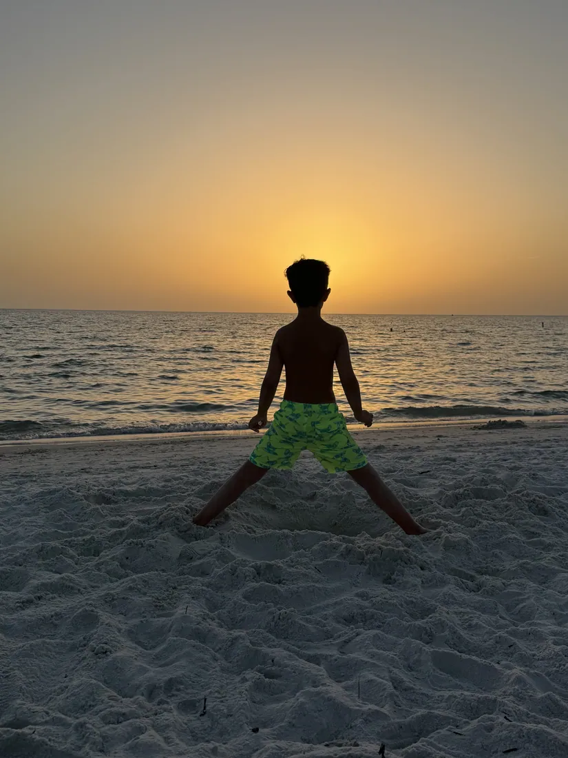 A kid planted in the sand at sunset, facing the water