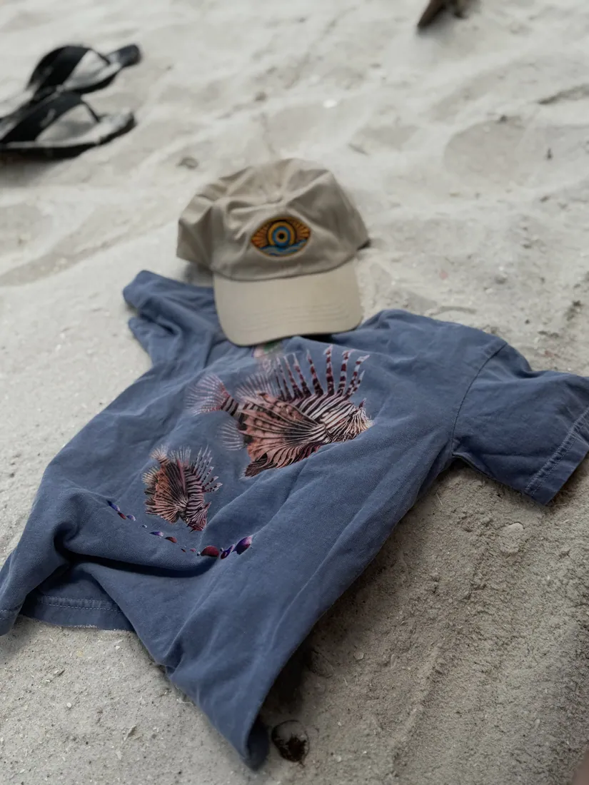 A lionfish tee and cap laid out on the sand